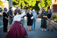 A woman in a nineteenth century costume points to a monument while an audience in masks looks on. It is on a footpath in Castlemaine with trees and shops visible