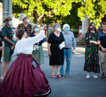 A woman in a nineteenth century costume points to a monument while an audience in masks looks on. It is on a footpath in Castlemaine with trees and shops visible